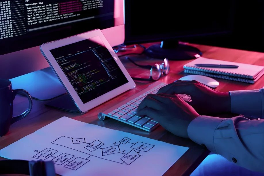 A person is typing on a white keyboard at a cluttered desk. A tablet displaying code, a monitor, and a cup are present. Nearby, paper with a flowchart, a notebook, and glasses are visible. The scene is lit with blue and pink tones, indicating a tech work environment.