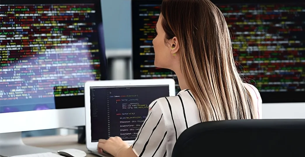 A woman with long, light brown hair sits at a desk using a laptop, surrounded by three large computer monitors filled with colorful lines of code. She appears focused on her work in a modern office setting.