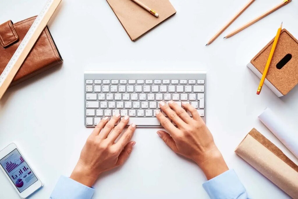 An aerial view of a workspace featuring a modern keyboard with hands typing on it. Surrounding the keyboard are various items including a brown leather wallet, a wooden ruler, several pencils, and a notepad. A smartphone displaying charts and graphs lies at the bottom left corner. The scene conveys a productive atmosphere, with clean lines and a minimalist aesthetic, focusing on the act of writing or data entry.