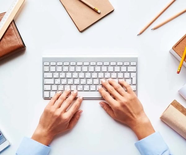 An aerial view of a workspace featuring a modern keyboard with hands typing on it. Surrounding the keyboard are various items including a brown leather wallet, a wooden ruler, several pencils, and a notepad. A smartphone displaying charts and graphs lies at the bottom left corner. The scene conveys a productive atmosphere, with clean lines and a minimalist aesthetic, focusing on the act of writing or data entry.